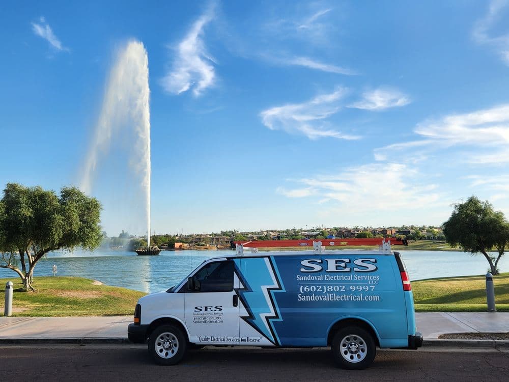 Blue service van "SES Sandoval Electrical Services" parked by a lake with a fountain.