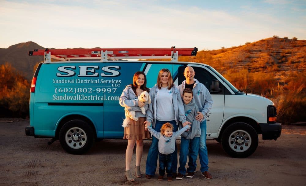Family with two children and a dog in front of a blue electrical services van at sunset.